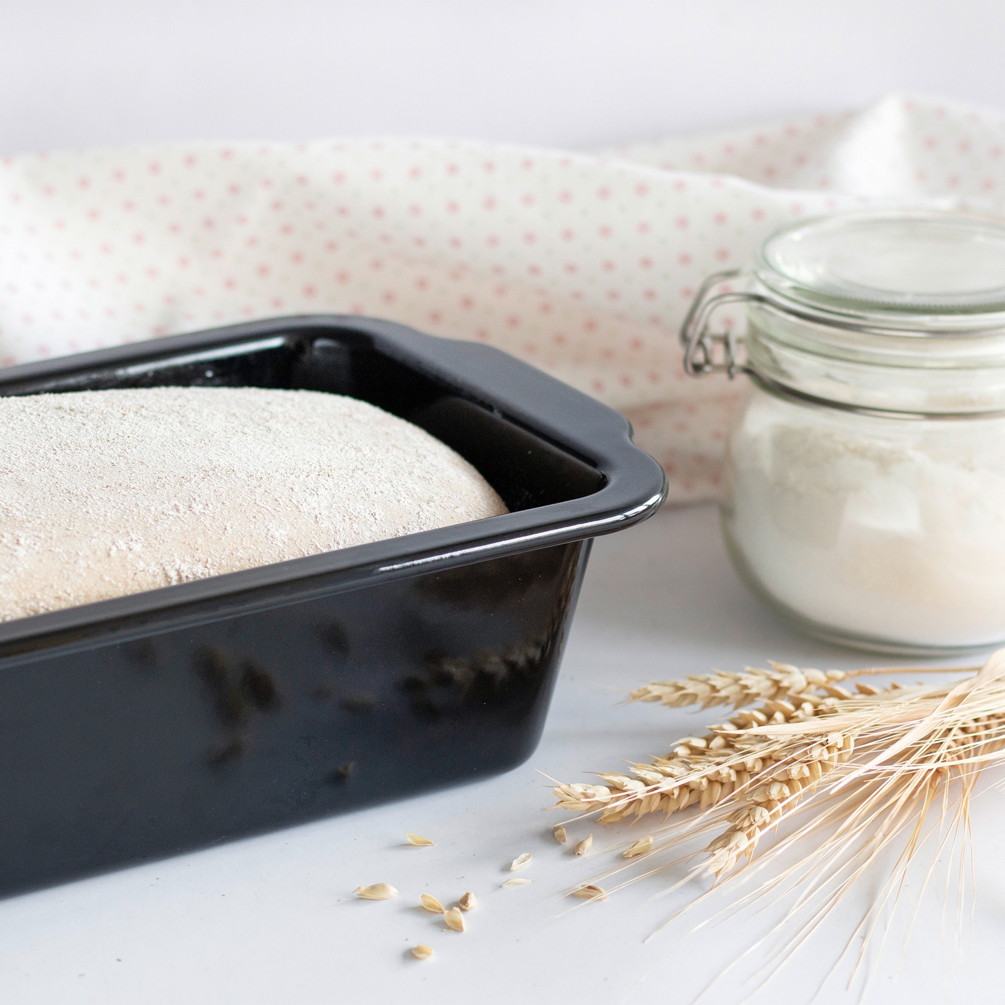 Brotteig in schwarzer Kastenform neben Mehlglas