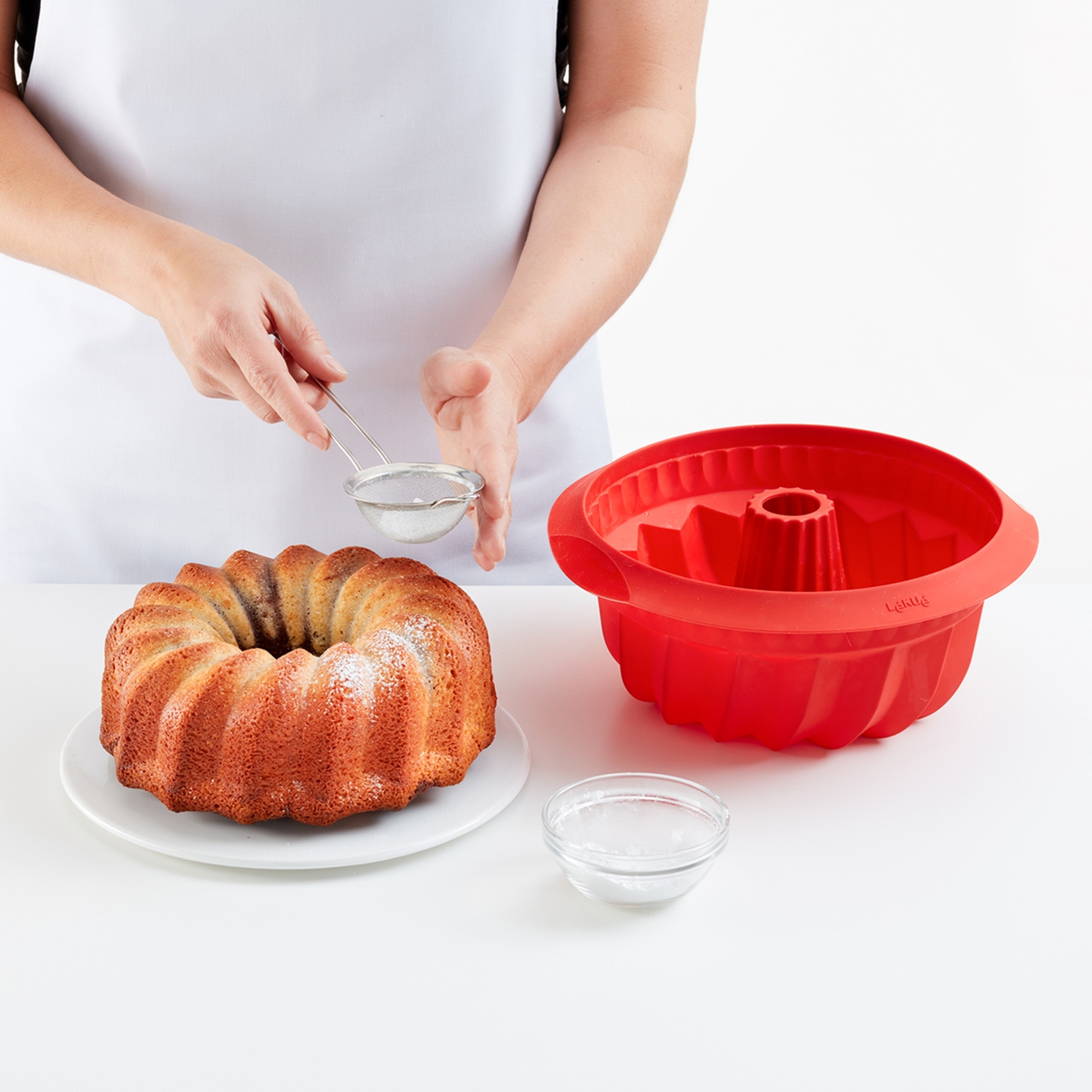 Person dusting bundt cake beside red silicone mould